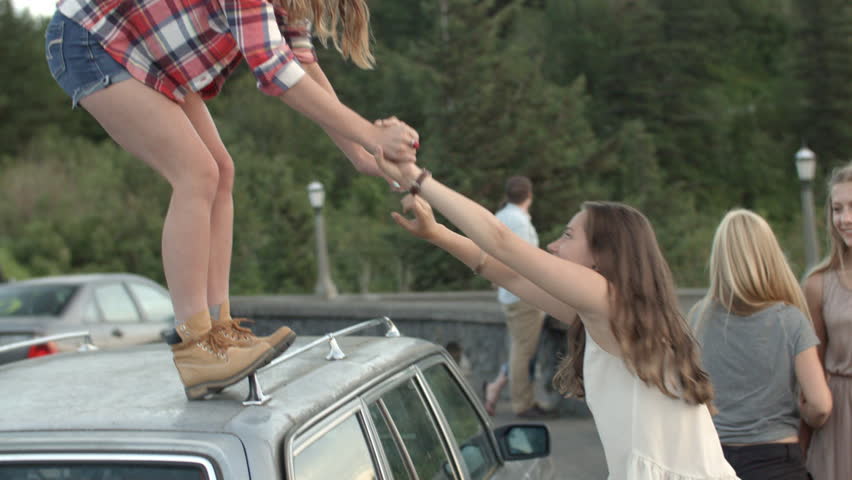 Girl Helps Her Friend Jump Off Of Car Roof They Hug And Laugh Stock